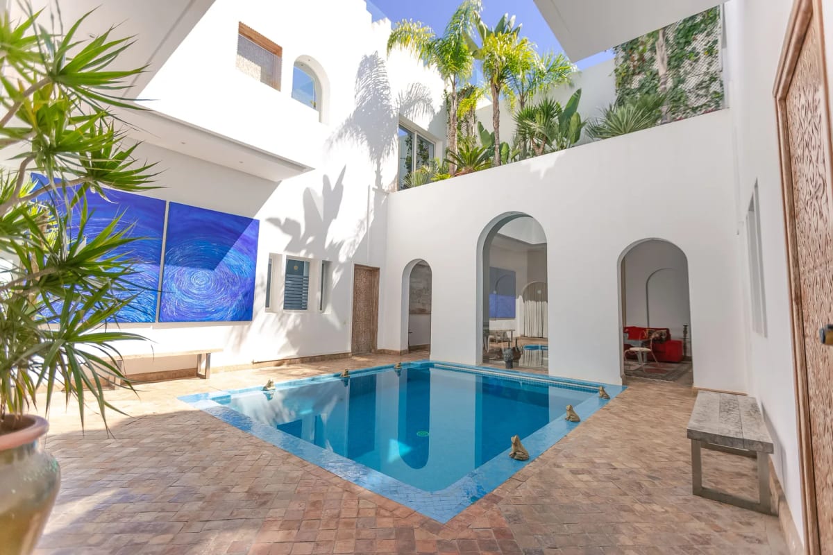 Patio d’une villa blanche marocaine avec piscine carrée, arcades ouvertes sur le salon et palmiers