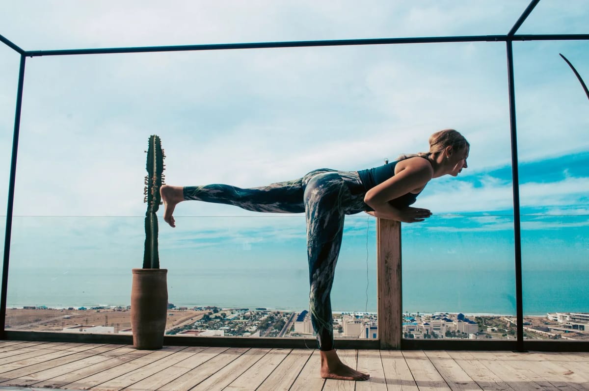Femme en posture de yoga sur une jambe, sur une terrasse en bois avec vue dégagée sur la mer