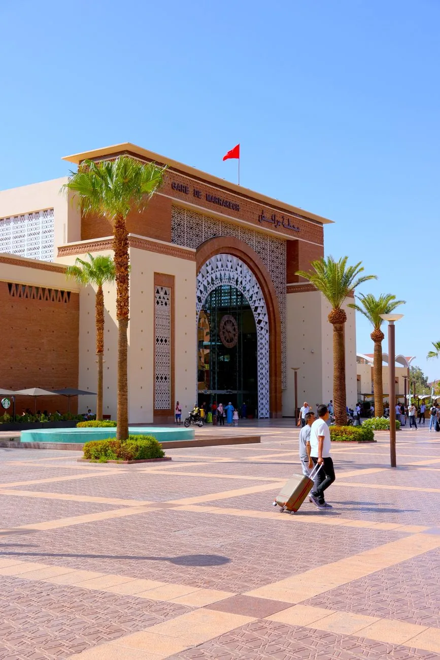 Gare ferroviaire marocaine en pierre ocre, palmiers et voyageurs sur le parvis